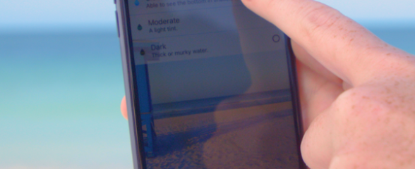 a black smartphone shows a screen with illegible text with a finger pointing at the screen. There is blue green ocean in the background and sandy beach